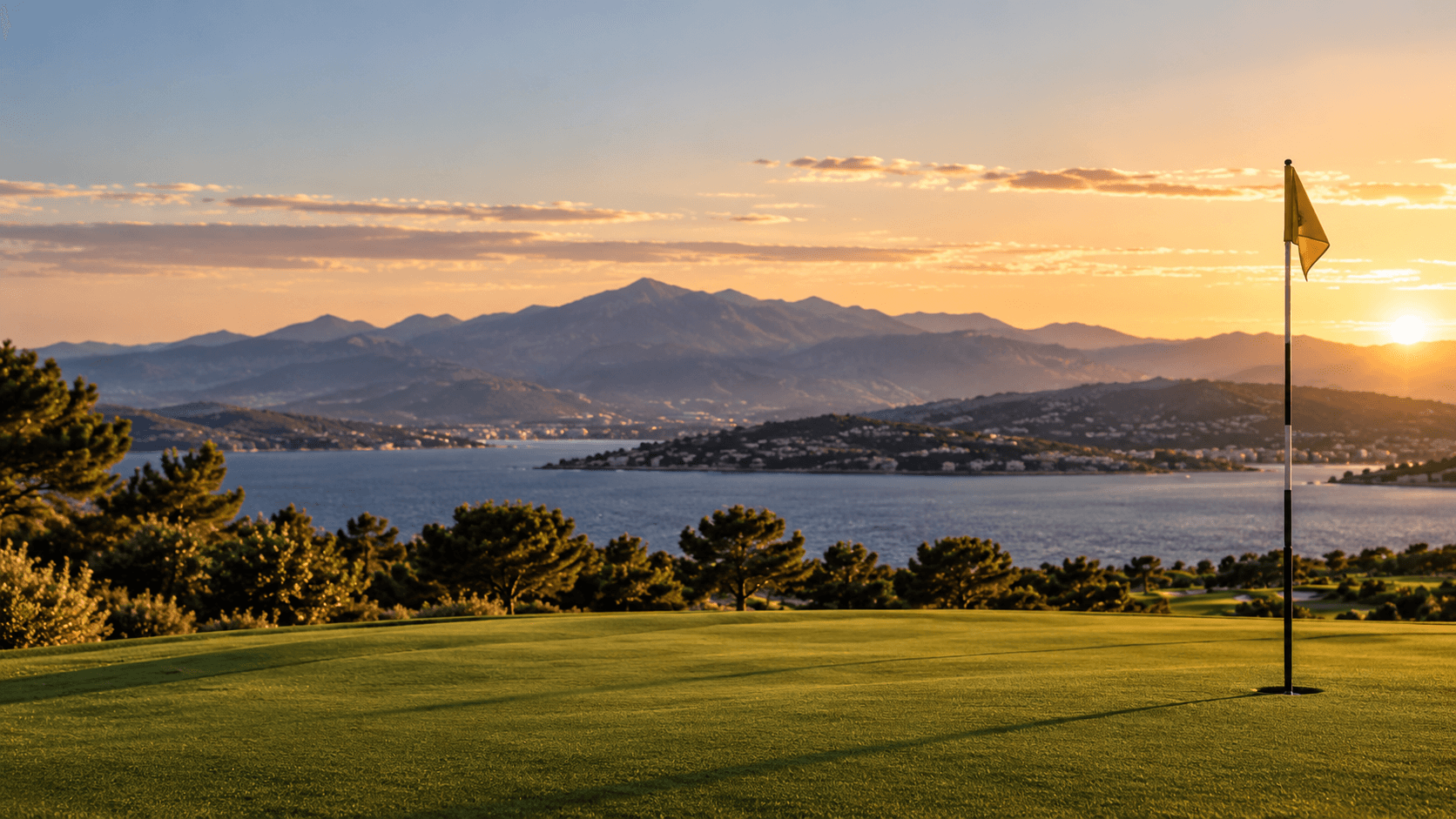 A flagstick on an immaculate green above the bay as the sun sets behind the mountains